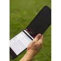 A scorebook is held in a hand with a pencil tucked inside it recording game statistics on a blank score sheet while surrounded by a grassy field.