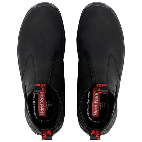 Black slip-on shoes are displayed from above showcasing a smooth exterior and elastic sides for easy wear with bold red branding visible inside the shoe indicating the brand name Hard Yakka.