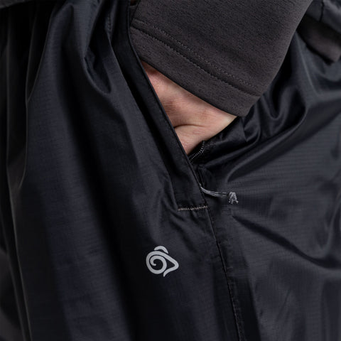 A hand is inserted into the side pocket of a black jacket with a logo on the lower part, showcasing the garment's design and functionality in a close-up view.