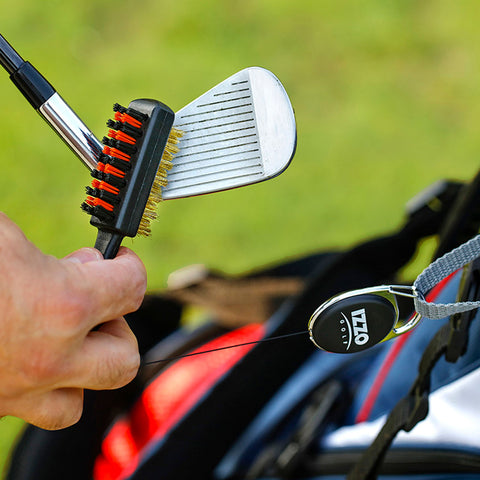 A hand holds a golf club cleaning brush against a golf club's head while a golf bag is visible in the background and grass covers the ground.