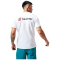 A person stands with their back to the viewer wearing a white t-shirt featuring a red square and the word "berghaus" in black on the back with blue shorts visible below.