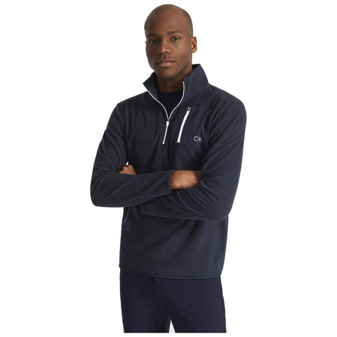 A man stands confidently with arms crossed wearing a dark fleece pullover featuring a zippered pocket and a logo on the chest in a plain white background setting.