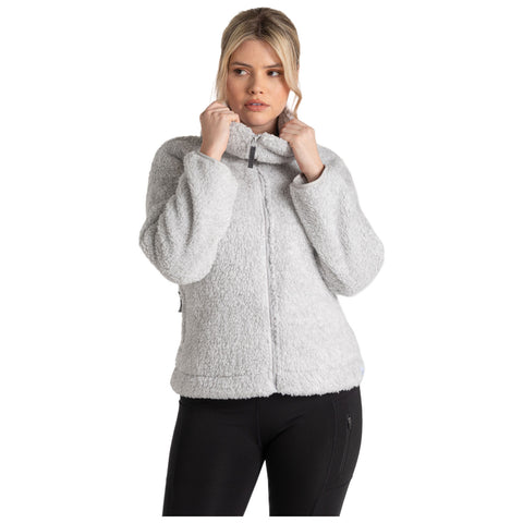 A woman is adjusting the collar of a light gray fleece jacket while standing against a plain white background, wearing black pants and displaying a thoughtful expression.