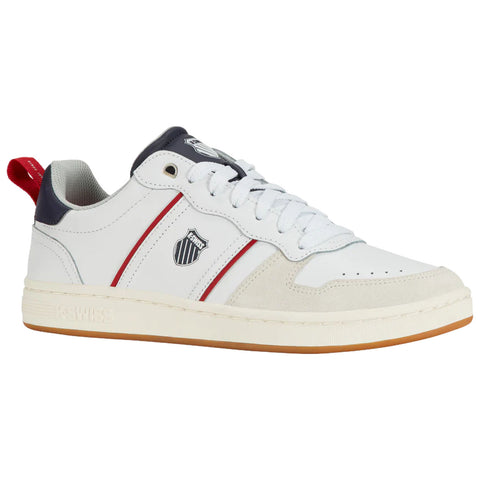 A sneaker rests on a flat surface showcasing a white leather body with navy and red accents along the sides featuring a shield logo and a gum rubber sole.