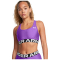 A person is wearing a purple athletic sports bra with a black band displaying the text "UNDER ARMOUR" while looking directly at the viewer in a neutral setting.