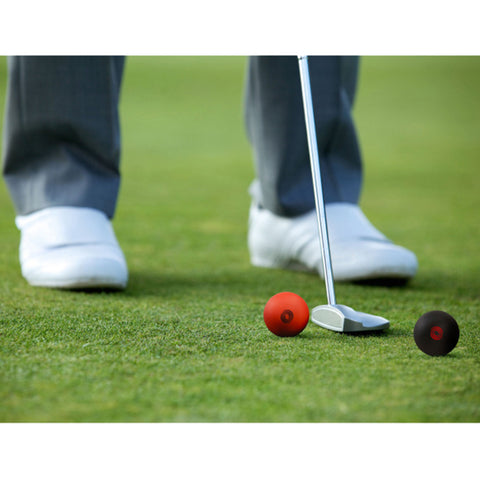 A golfer in a gray suit and white shoes prepares to putt a red ball on green grass with a putter while a black ball rests nearby.