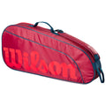 A red sports bag with a prominent orange Wilson logo sits upright displaying its elongated shape and a blue handle on top for easy carrying.