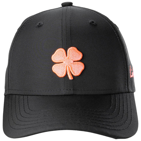 A black baseball cap displays a bright orange four-leaf clover emblem on the front while featuring a structured brim and stitched detailing throughout the fabric