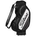 A black and white golf bag stands upright with compartments for clubs and a padded shoulder strap for carrying in a golfing environment. The logo prominently displays "Titleist."