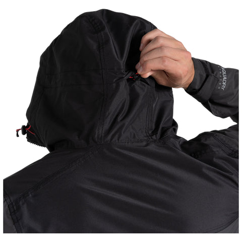 A person adjusts the drawstring of a black hooded jacket with one hand while the other hand is positioned by the hood in a light environment.