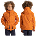 A child wearing an orange jacket stands confidently with hands in pockets the background is plain white highlighting the jacket's color and the child's curly hair.