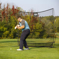 A golfer is swinging a club at a white ball on a green field surrounded by trees, with a protective net behind him catching stray shots.