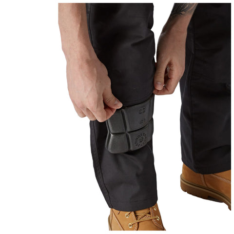 A hand secures a knee pad onto the leg of dark work pants while wearing a tan boot. The setting suggests preparation for a physically demanding task.