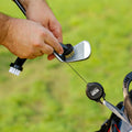 A person is cleaning a golf club head with a brush while holding a retractable cleaning tool attached to their golf bag in a grassy outdoor setting.