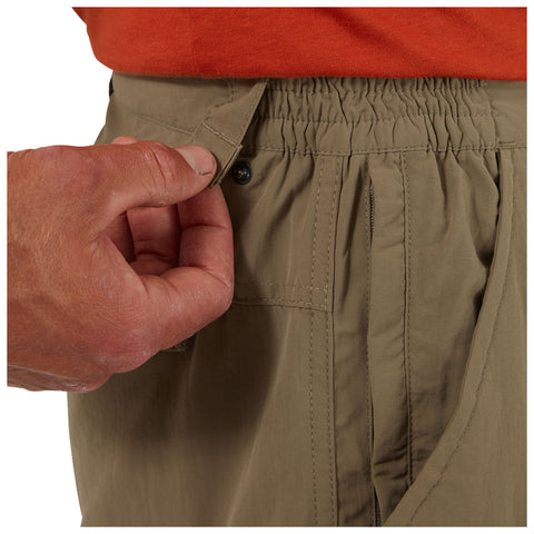 A hand is fastening a button on a pair of beige pants with an elastic waistband the individual is wearing an orange shirt in a neutral background.