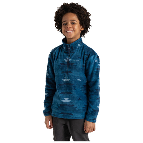 A young boy with curly hair wears a blue patterned fleece pullover and smiles while standing in a neutral background, exuding a cheerful and relaxed demeanor.