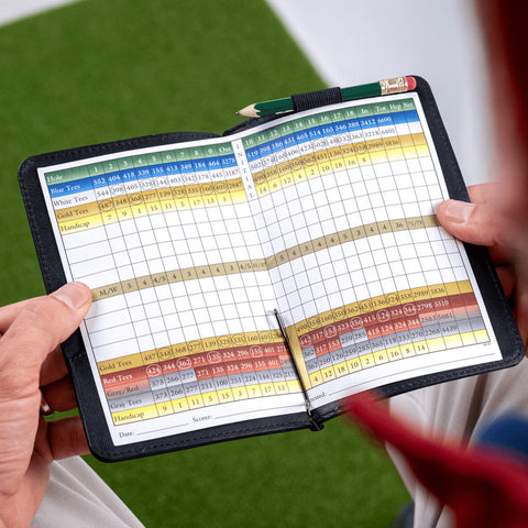 A hand holds an open golf scorecard displaying holes distances and scoring options arranged in a grid format while resting on a green surface suggesting a golf course environment.