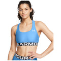 A female model is wearing a blue sports bra with a black and white logo band. She stands confidently with her hand on her hip against a plain background.