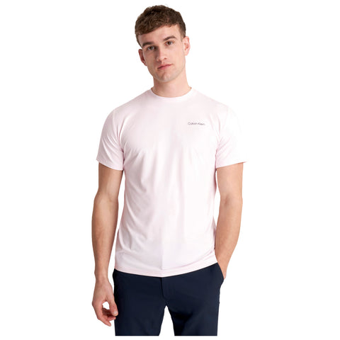 A young man stands confidently wearing a light pink Calvin Klein t-shirt and dark pants with one hand in his pocket in a plain white background.