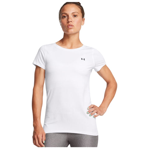 A woman stands confidently wearing a fitted white athletic shirt with a small black logo. She has her left hand on her hip and a neutral expression in a plain background.