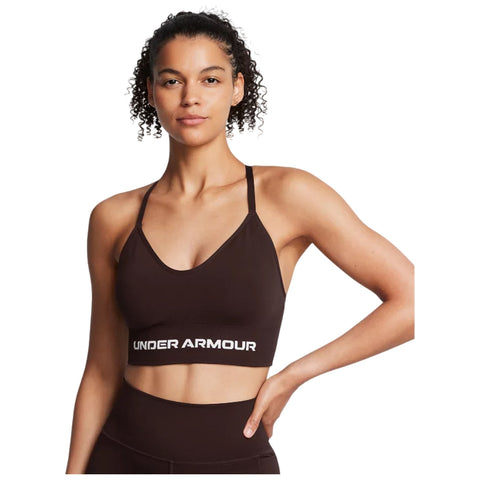 A woman is standing confidently wearing a brown athletic crop top branded with Under Armour she has curly hair and a neutral expression against a plain white background.