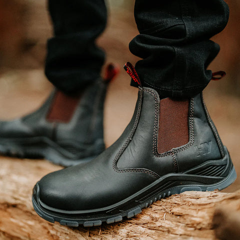 A pair of black leather boots with elastic side panels are positioned on a log in a forested area, surrounded by blurred natural elements, suggesting an outdoor setting.