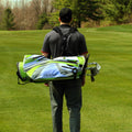 A person is carrying a green and blue golf bag on their back while walking across a grassy golf course surrounded by trees in the background.