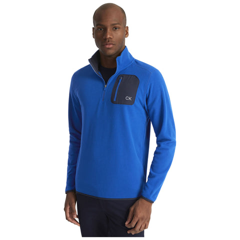 A man stands wearing a bright blue fleece pullover with a zippered collar and a black chest pocket featuring the initials CK, showcasing a casual style in a neutral background.