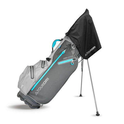 A gray golf bag with turquoise accents stands upright on thin legs featuring a black rain cover extended over the top protecting the contents from moisture in an outdoor setting.