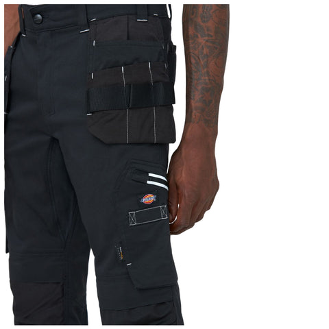 A close-up of a person's black work pants features multiple pockets and white stitching. The person's hand is positioned near the pockets, highlighting the functional design for work-related tasks.