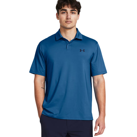 A young man stands confidently wearing a blue polo shirt with a logo on the left chest his hands are casually placed in his pockets in a neutral background.