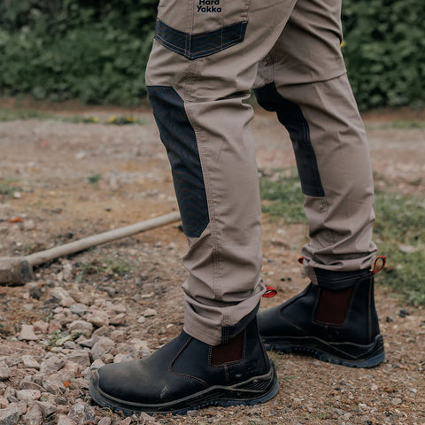 Tan work pants with dark patches are worn by a person standing on a gravel surface wearing black boots while a shovel leans against the ground in a natural outdoor setting.