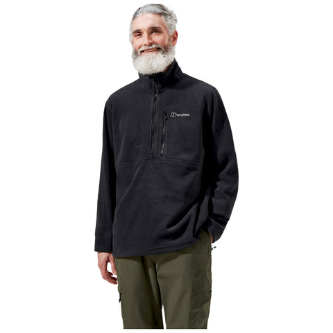 A man with a beard smiles while wearing a black fleece pullover with a zippered pocket on the chest and green cargo pants standing against a white background.