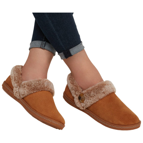 Brown slippers with a soft, fuzzy lining rest on crossed legs wearing dark denim jeans, showcasing a relaxed and cozy style in a casual indoor environment.