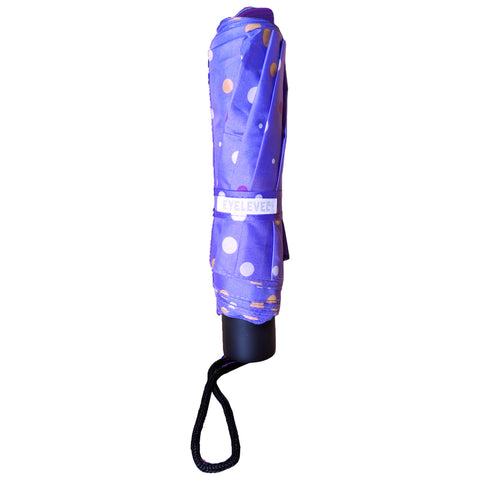 A purple umbrella is rolled up and secured with a strap featuring polka dots in various colors its handle has a black strap for easy carrying in an outdoor setting