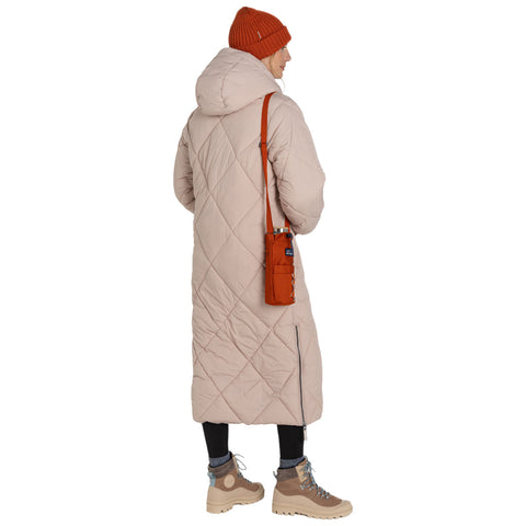 A person wearing a long light beige quilted coat and a knitted orange beanie is standing with their back turned, carrying a small orange bag, and wearing tan boots in a neutral setting.