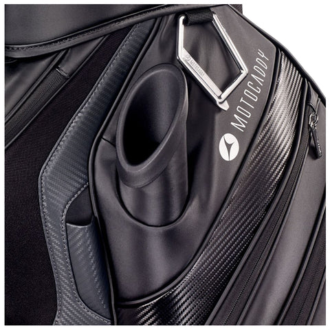 A black golf bag displays a cylindrical holder for alignment sticks alongside a silver carabiner and a zippered pocket with the brand name Motocaddy prominently shown.