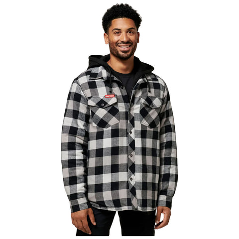 A man stands smiling wearing a black and white checkered flannel shirt over a black hoodie in a neutral background emphasizing his outfit and expression.