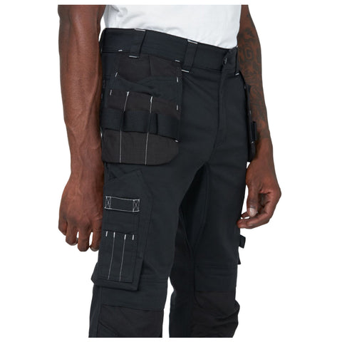 Black cargo pants fitted with multiple pockets and straps are being worn by a person standing against a plain background showcasing a close-up view of the pants and a partially visible hand.