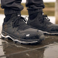 Black athletic shoes with a textured surface stand on a wet surface reflecting water. The laces are tied, and some dirt is visible on the shoes, suggesting outdoor use.