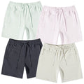 Four pairs of shorts in soft fabric feature an elastic waistband with drawstrings. They are displayed in pastel green, light pink, dark gray, and cream colors against a plain background.