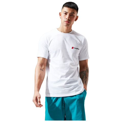 A man stands in a white t-shirt and turquoise shorts with a confident expression his right arm raised slightly displaying a tattoo in a minimalistic, bright background.