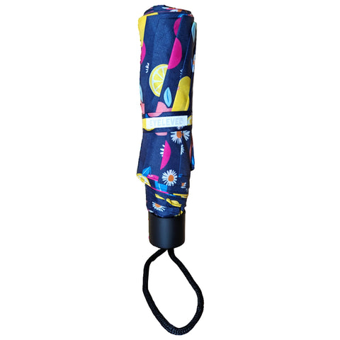 A closed umbrella features a colorful fabric design with fruit and floral patterns resting vertically with a black handle and a matching strap for easy carrying.