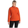 A woman wearing an orange fleece pullover stands with a serious expression. The pullover features a half-zip and is paired with black leggings, creating a casual athletic look.