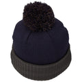 A knitted beanie with a dark blue top and a green ribbed cuff sits upright showcasing a fluffy pom-pom on top in a neutral setting suitable for winter wear.