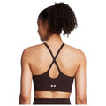 A woman with curly hair is wearing a dark brown athletic sports bra featuring crisscross straps on the back standing against a plain background emphasizing the clothing design.