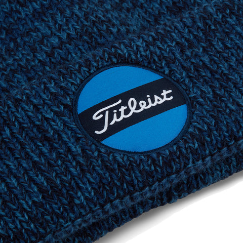 A blue knitted beanie displays a circular patch featuring a blue background with a black stripe and the text Titleist in white script, resting on a textured surface.