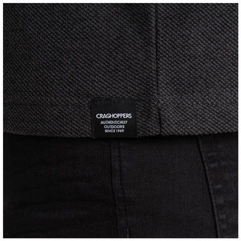 A dark textured fabric features a visible clothing label while being worn above black pants in a neutral setting emphasizing the outdoor brand Craghoppers which promotes authentic outdoor wear since 1965.