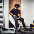 A man sitting on a staircase is examining a dark tool with a red label while dressed in work attire surrounded by tools and equipment in a slightly cluttered interior space.