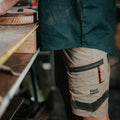 A person stands at a workbench touching a wooden piece wearing dark clothing and beige shorts with pockets and branding in a workshop filled with tools and materials.